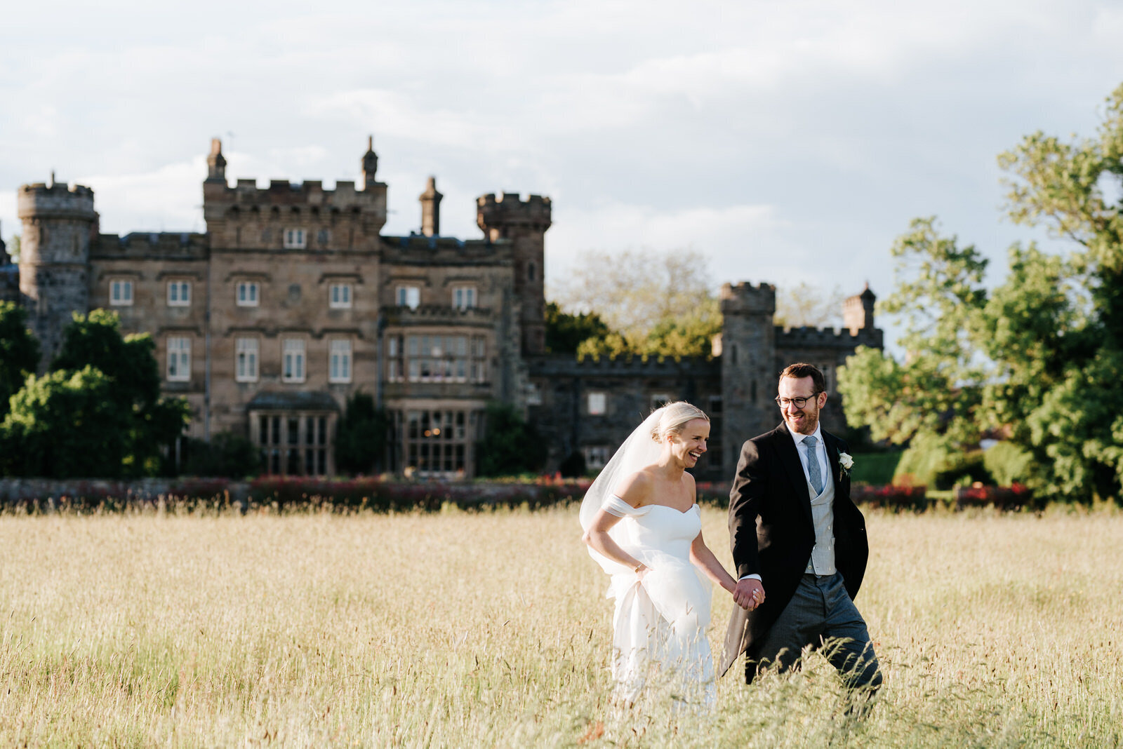 Bride and groom walk through lush fields towards marquee as Hawarden Castle is in the background