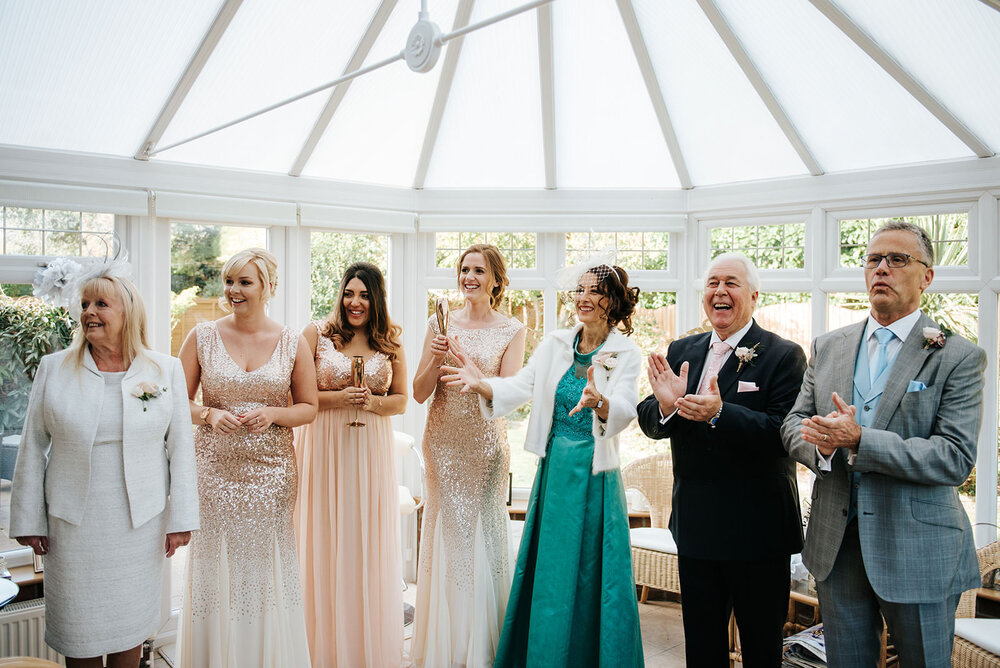 Parents of the bride and mother of the groom clap and cheer as they stand in conservatory and bride comes down the stairs, off-frame
