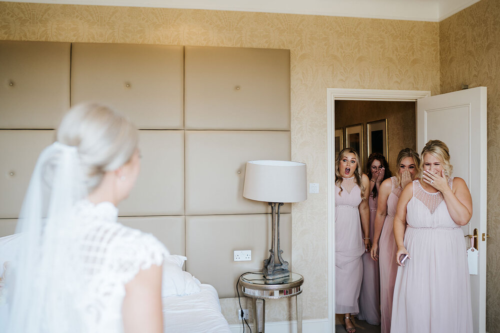 Bridesmaids in pink dresses react with joy and awe to seeing the bride in her dress for the very first time