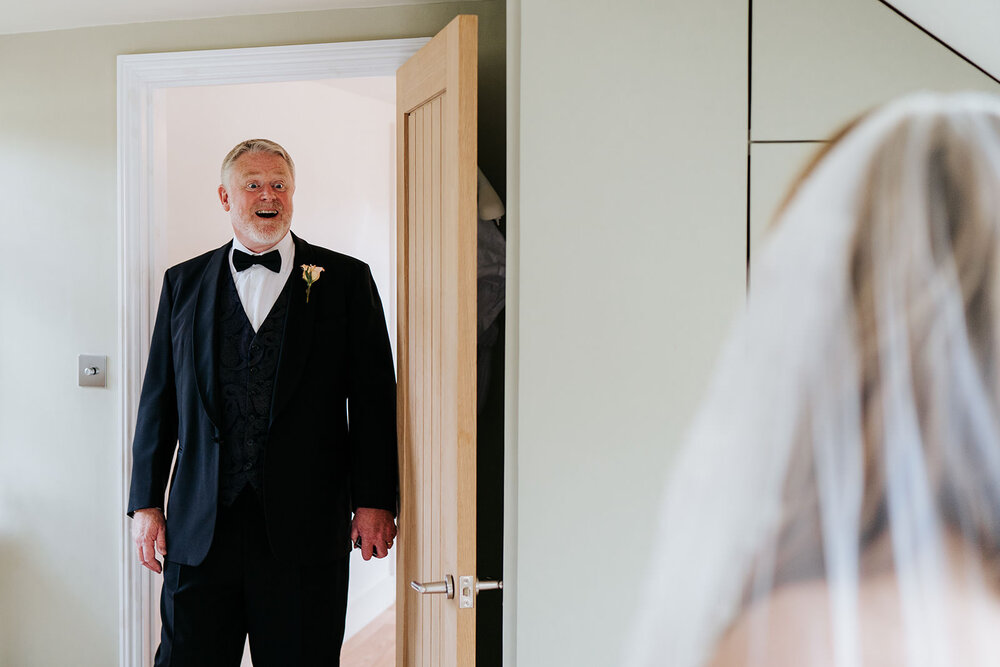 Father of the bride walks through doorway with mouth open as he sees his daughter in her wedding dress for the first time