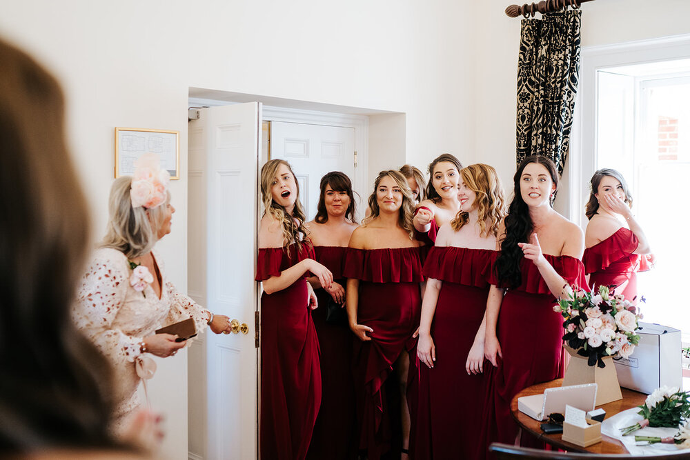 Bridesmaids in red dresses react with joy and awe to seeing the bride in her dress for the very first time