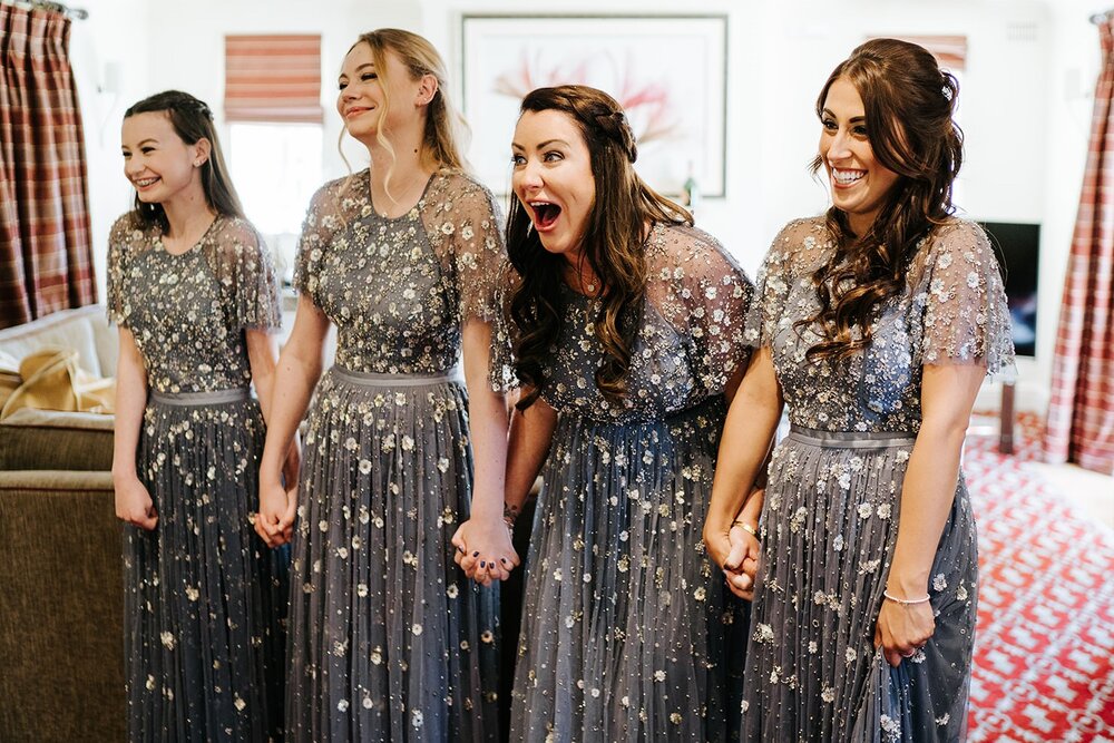 Bridesmaids hold hands and cannot contain their excitement as they see the bride, off-frame, in her dress for the very first time