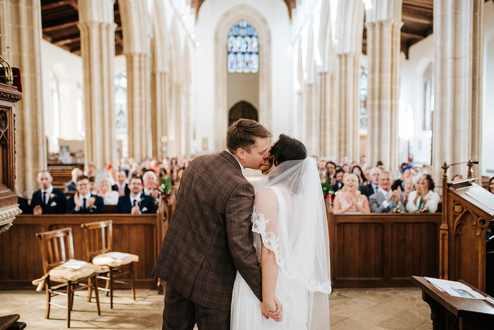 Wide photograph of church and guests as bride and groom hold hands and kiss in the foreground during their wedding ceremony