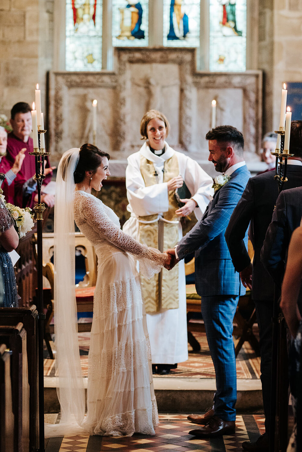 Bride holds groom's hands and leans back with mouth open as she cannot contain her excitement after the first kiss during their religious wedding ceremony in the Cotswolds