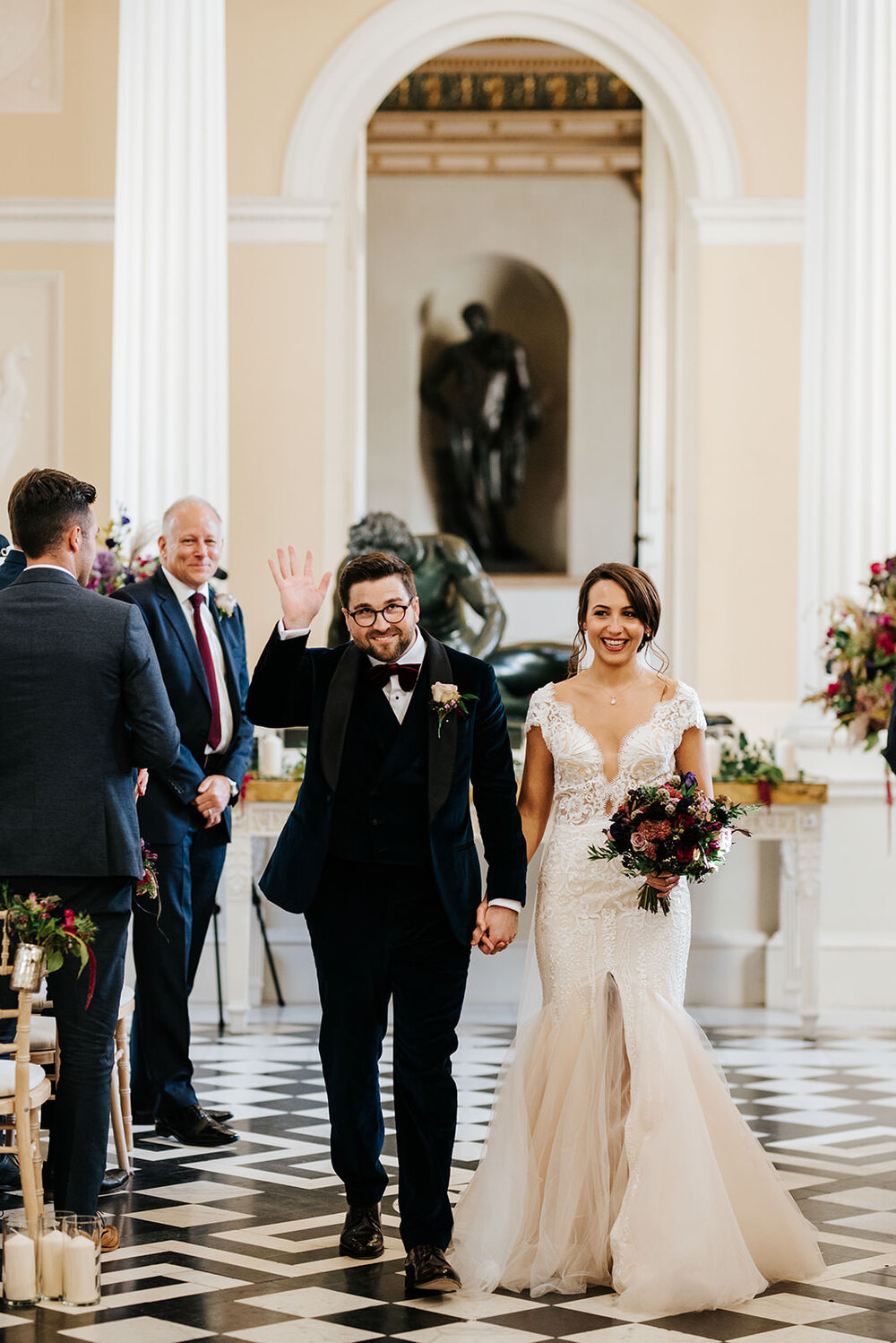 Bride and groom walk back down the aisle as married couple while groom waves at guests during Syon Park wedding