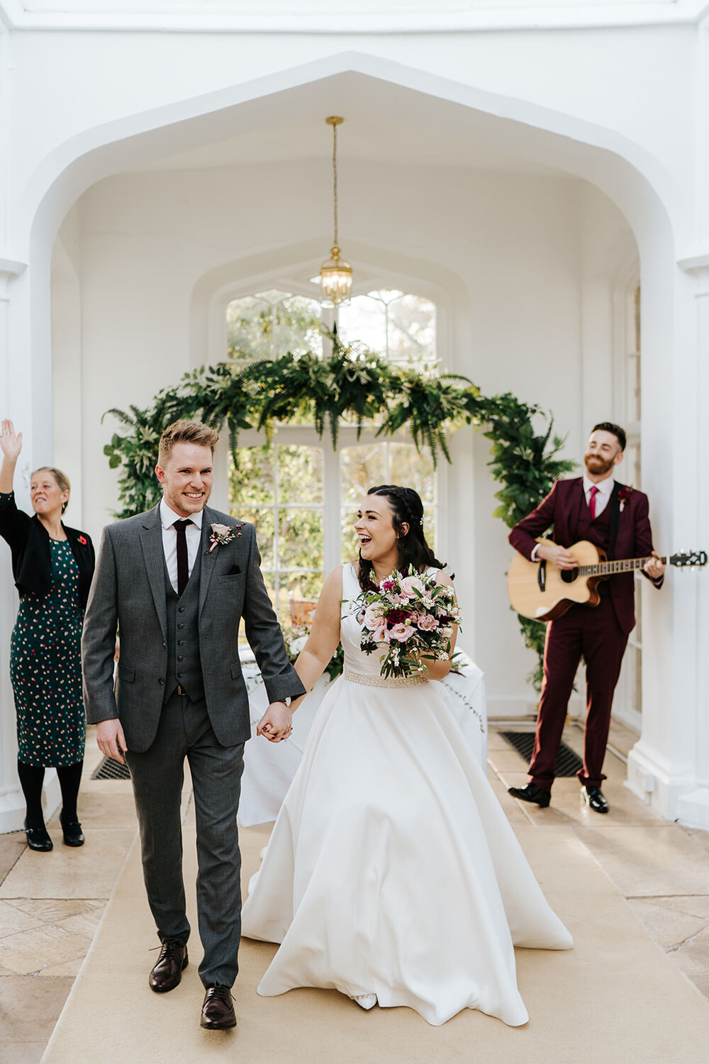 Bride and groom walk back down the aisle as married couple while friend, in the background, plays their favourite song on guitar