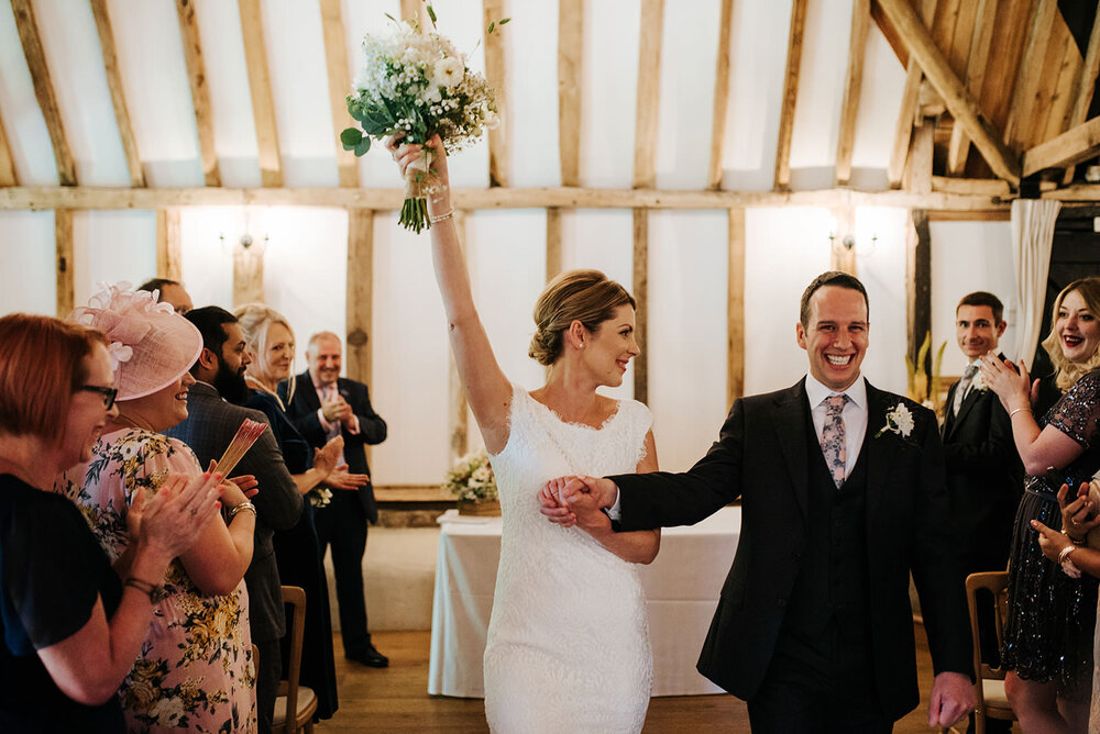 Bride lifts her bouquet into the air as she starts walking back down the aisle holding her husband's hand in barn wedding