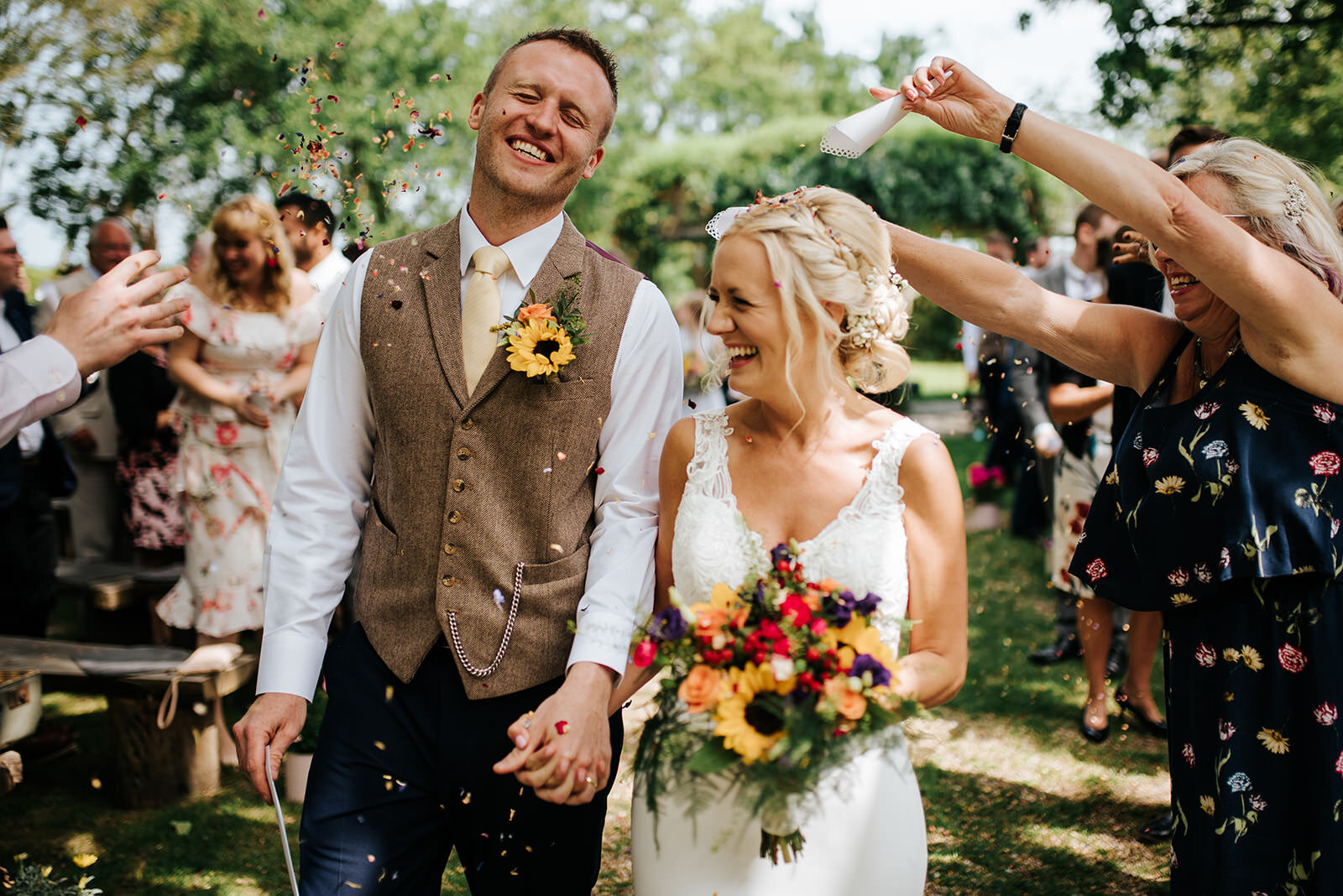 Bride and groom walk down the aisle as guests throw confetti at them. Groom smiles as he nearly avoids getting confetti in the face. 