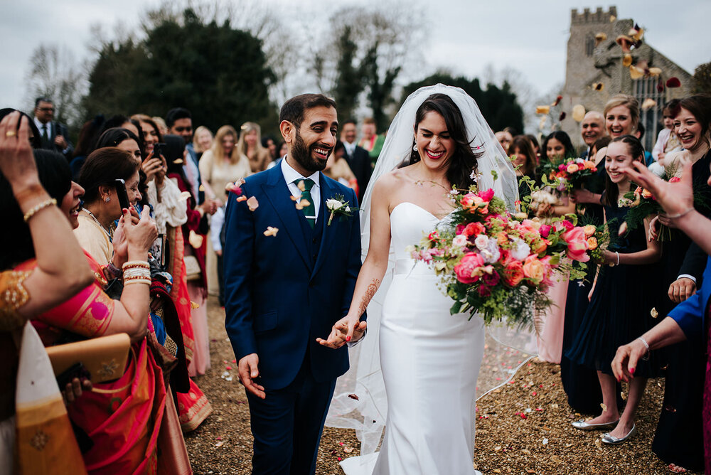 Bride and groom look surprised and happy at how much confetti is thrown at them after religious wedding ceremony in England