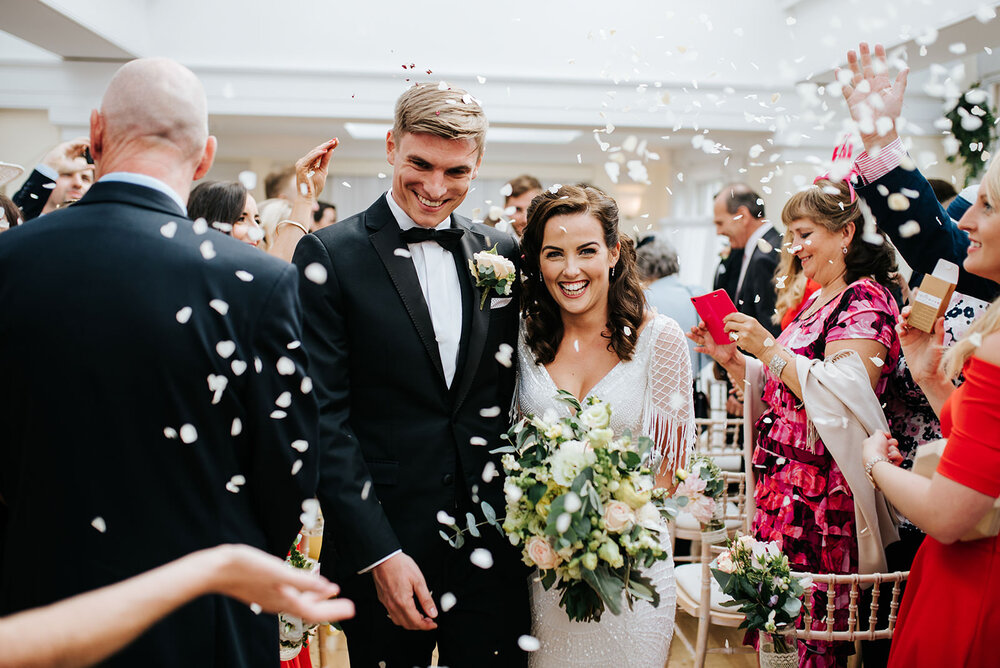 Bride and groom walk back down the aisle in Pembroke Lodge in Richmond and smile as guests throw white flower petal confetti at them 