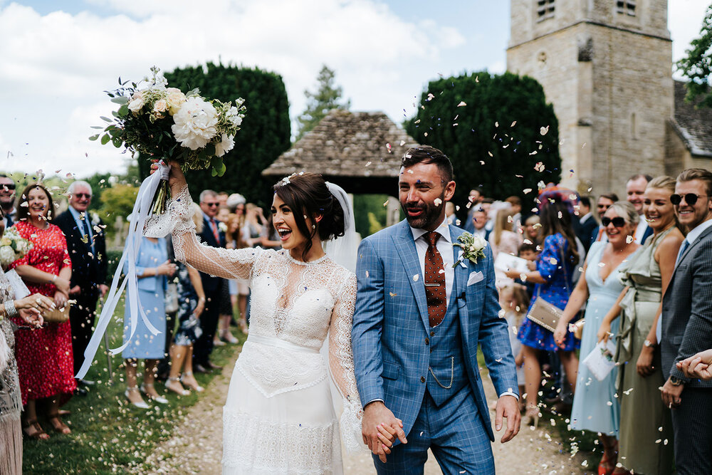 Bride and groom look joyful after wedding ceremony in Cotswolds, England. Bride lifts her bouquet in elation as groom receives handful of confetti to the face. 
