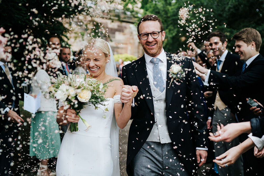 Bride and groom walk through tunnel of guests as they get pelted with confetti from both sides in wedding in Hawarden, Wales. 