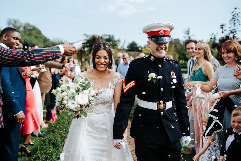 Bride and groom walk through tunnel of guests as they are attacked with confetti. Groom clenches jaw as impending fistful of confetti approaches. 