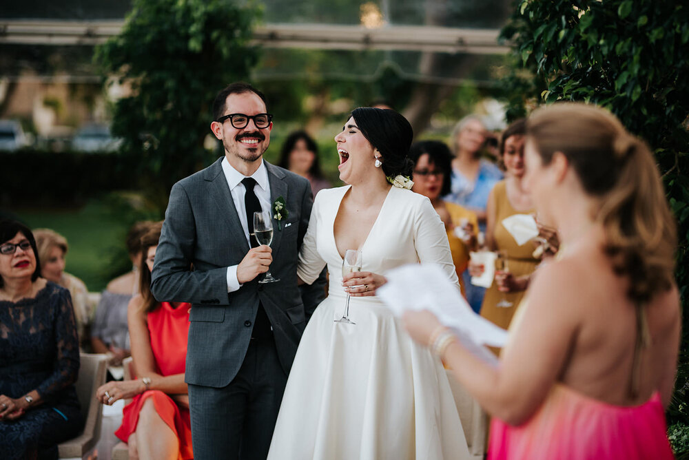 Bride and groom hold each other by the waist and laugh out loud as friend, in foreground and dressed in pink dress, reads her wedding speech