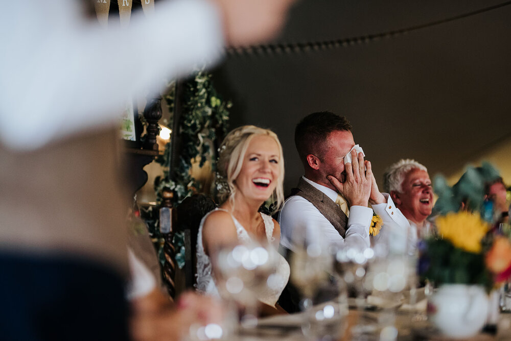 Best man, out of focus, reads his wedding speech as groom covers his face in embarrassment and bride smiles at best man