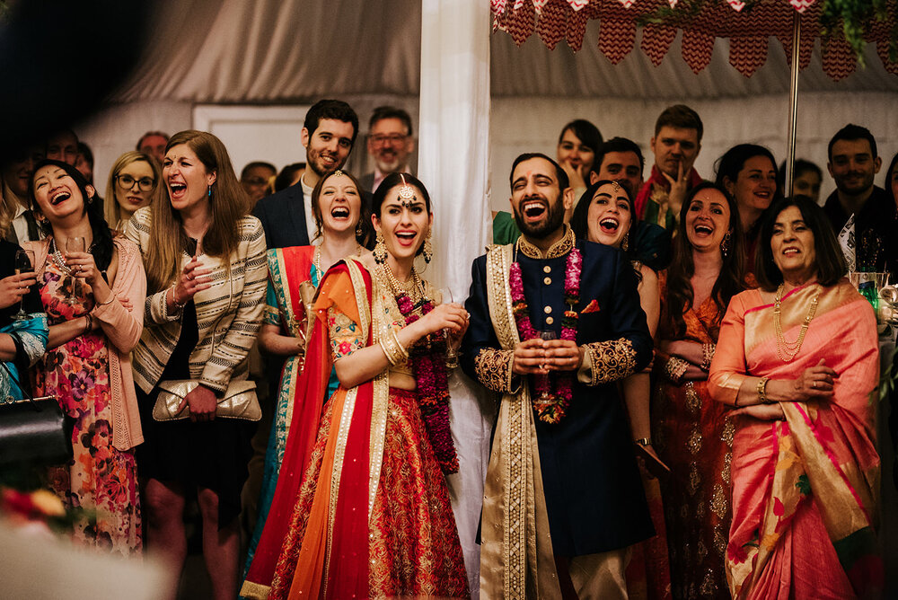 Bride and groom, in traditional Hindu dress, cannot contain the laughter as one of their friends, out of the frame, delivers their wedding speech. 