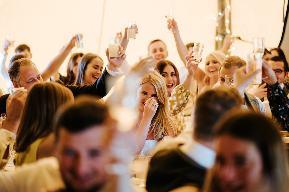 Bride, in the middle of the frame, wipes tears off her cheeks as all guests sat around her lift their glasses during wedding speeches