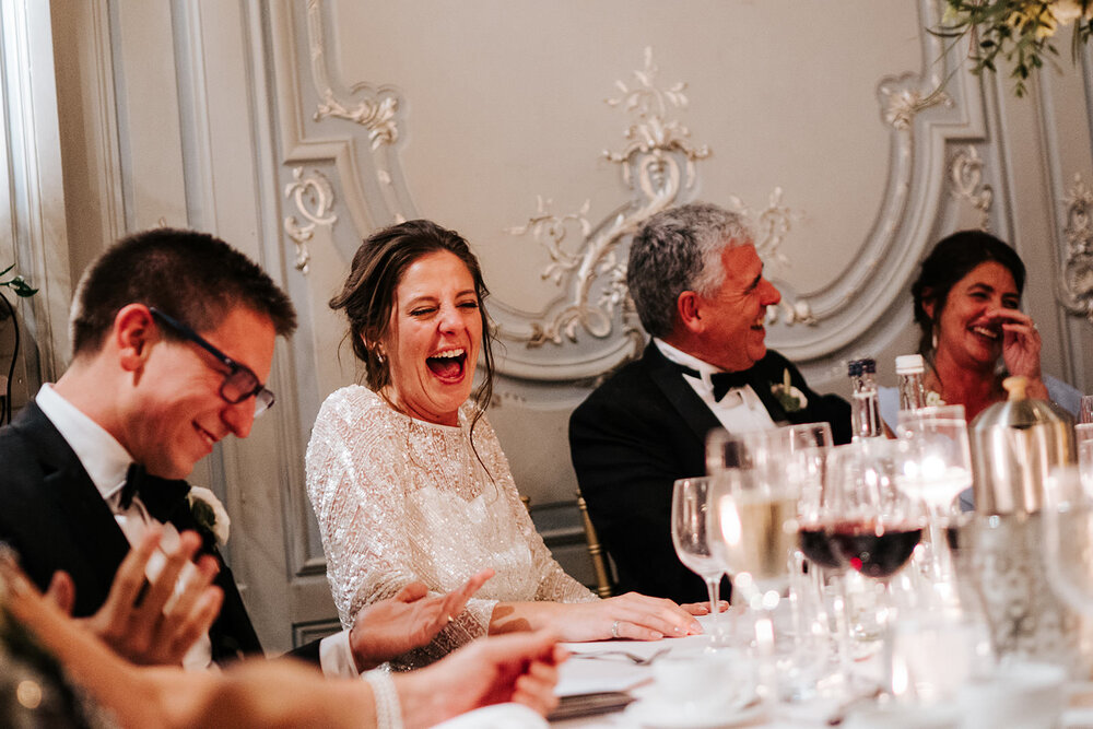 Groom, bride and parents of the bride laugh hysterically as speech is delivered off-frame during wedding at Savile Club in London