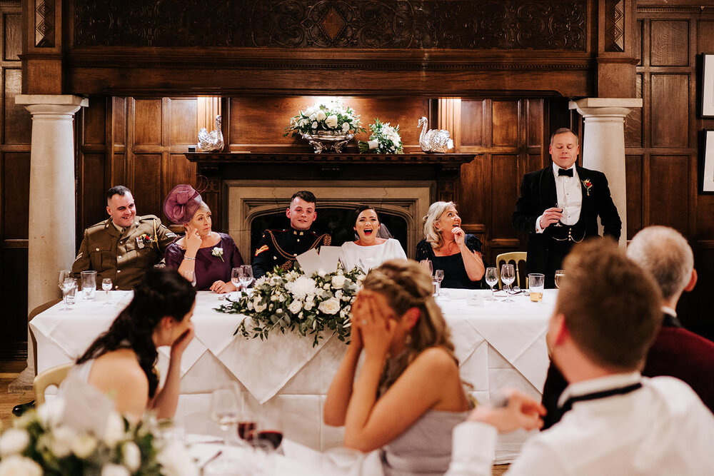 Wide photograph of the head table during wedding speeches while guests and family members react with laughter and embarrassment to what is being said