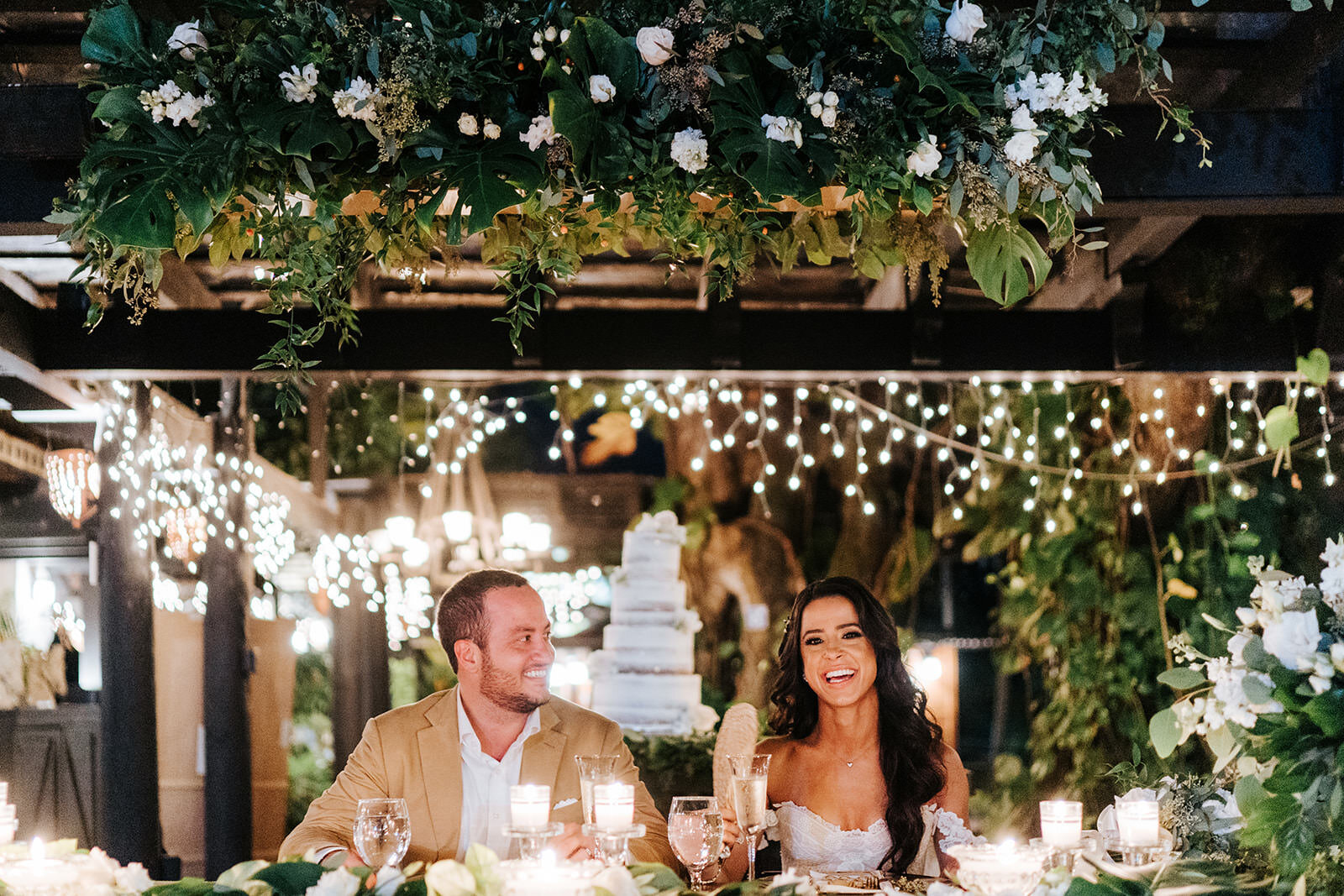 Bride and groom, framed by tropical foliage and wooden arches, smile and look at each other while wedding speech is delivered off-frame during wedding in Puerto Rican Mountain Hacienda
