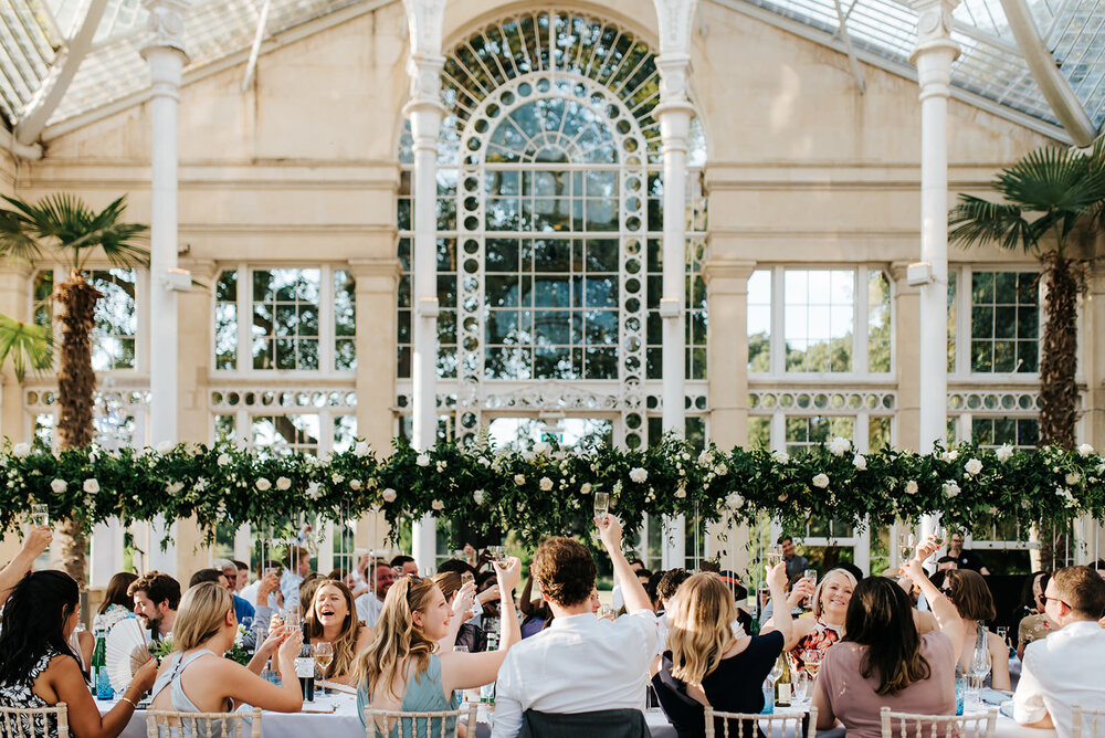 Wide photograph of guests raising a toast during wedding at Syon Park in London while flower arch by Emma Soulsby graces the middle of the frame