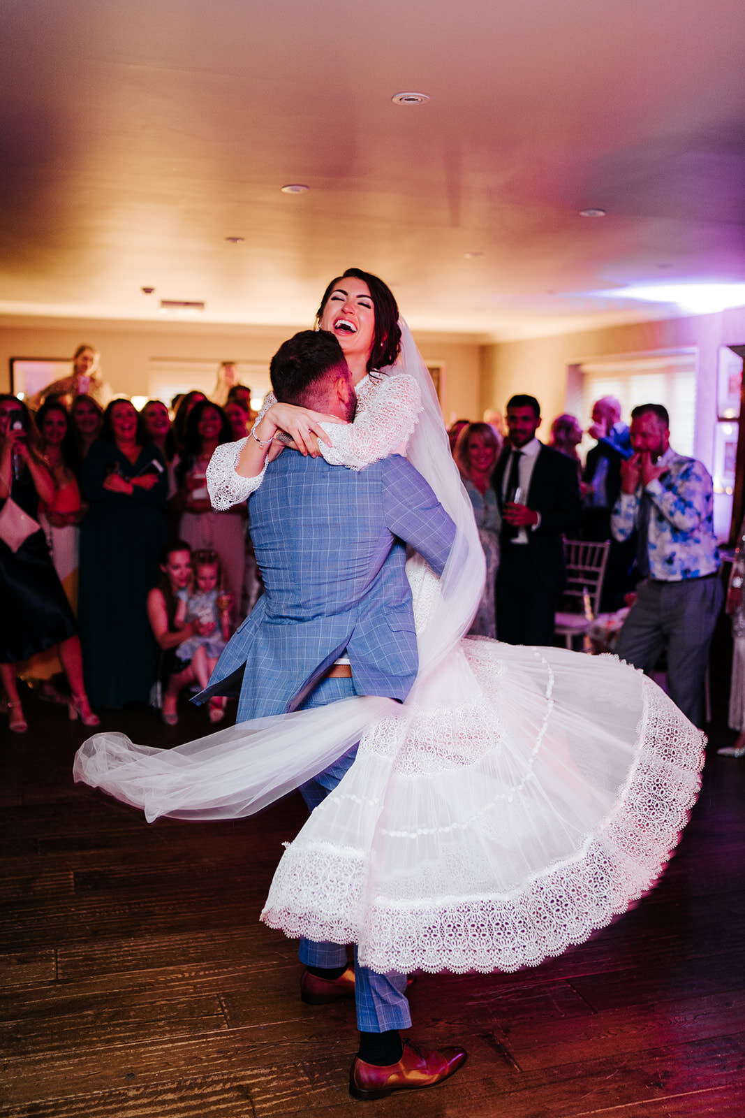 Groom picks up bride and swirls her around during first dance at Cotswolds wedding. Bride's dress trails along beautifully as groom turns her.