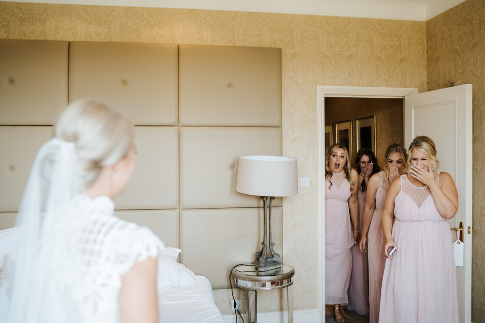 Bride stands out of focus in foregrounds as bridesmaids enter the room and see her in her wedding dress for the first time