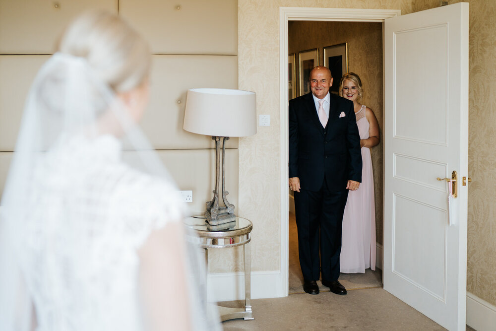 Father of the bride stands in the frame of the door as he sees her in her dress for the very first time