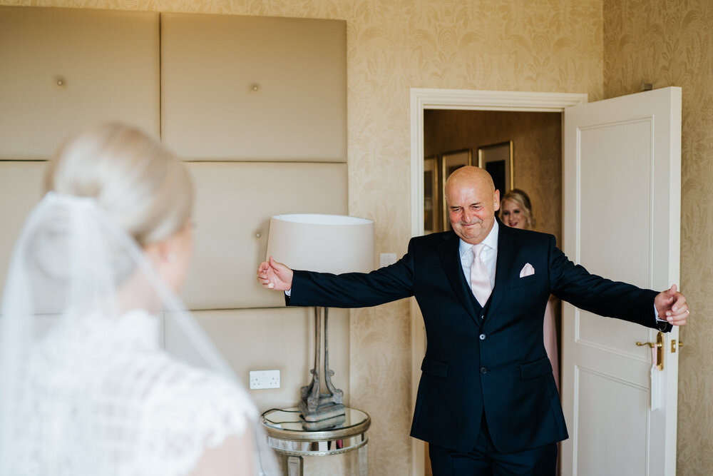 Father of the bride enters the room and opens both arms wide and cannot contain his excitement at seeing his daughter in her wedding dress