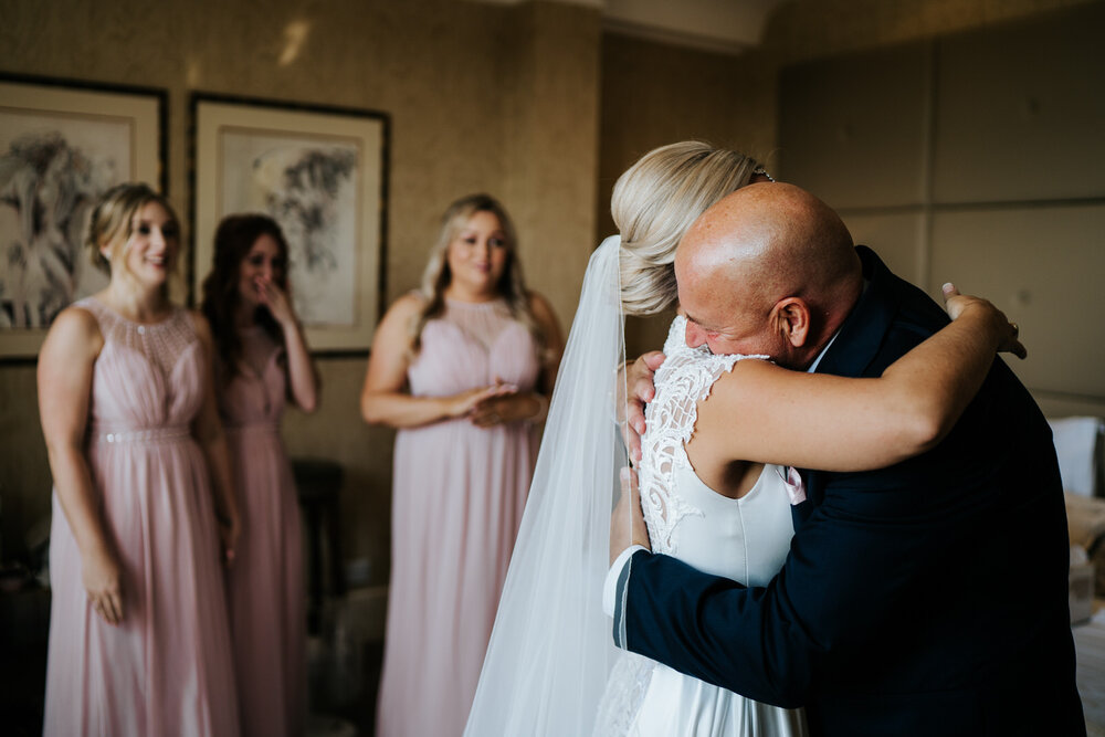 Father of the bride and bride hug in the foreground as bridesmaids look emotionally from the background at The Petersham Hotel