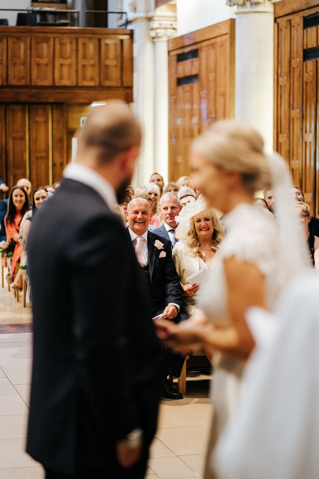 Bride and groom, blurred in the foreground, look towards their guests and make eye contact with the bride's parents, in focus, who smile at them