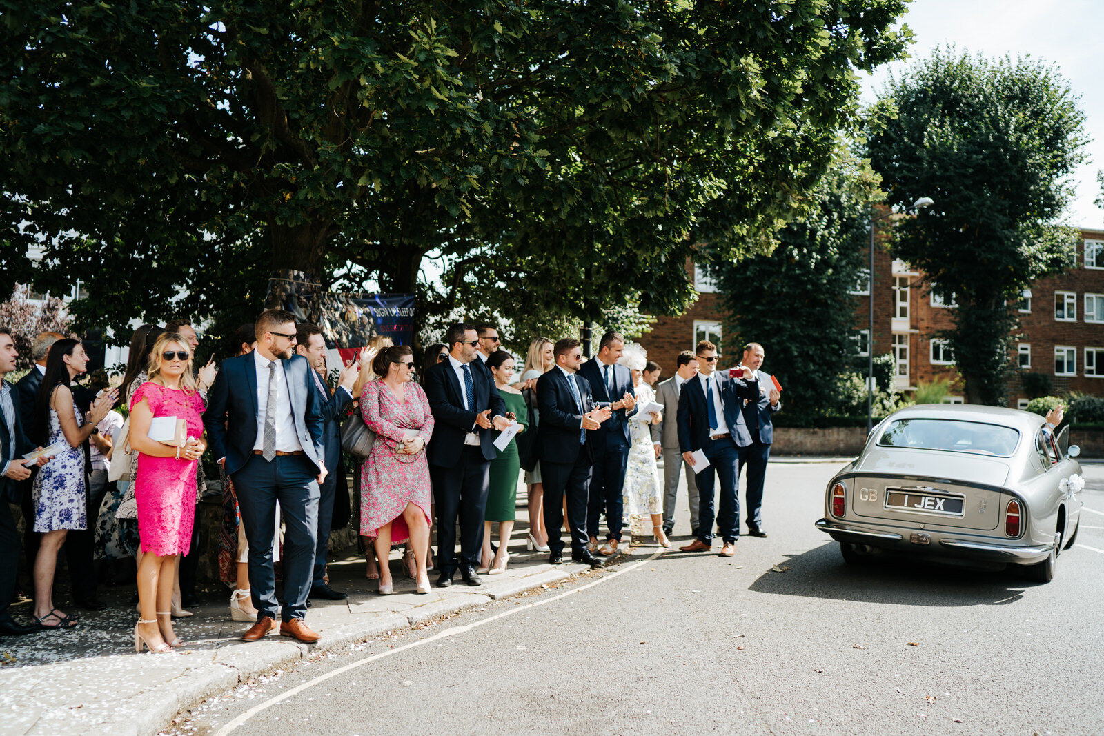 Guests stand on the left and wave as couple depart the church in vintage wedding car on the right