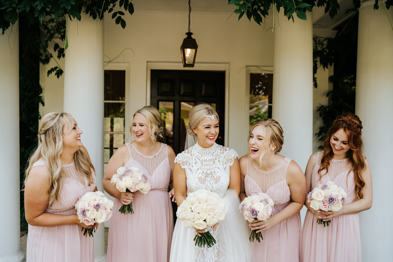 Bride and bridesmaids stand under lush foliage at entrance of Pembroke Lodge and smile at each other