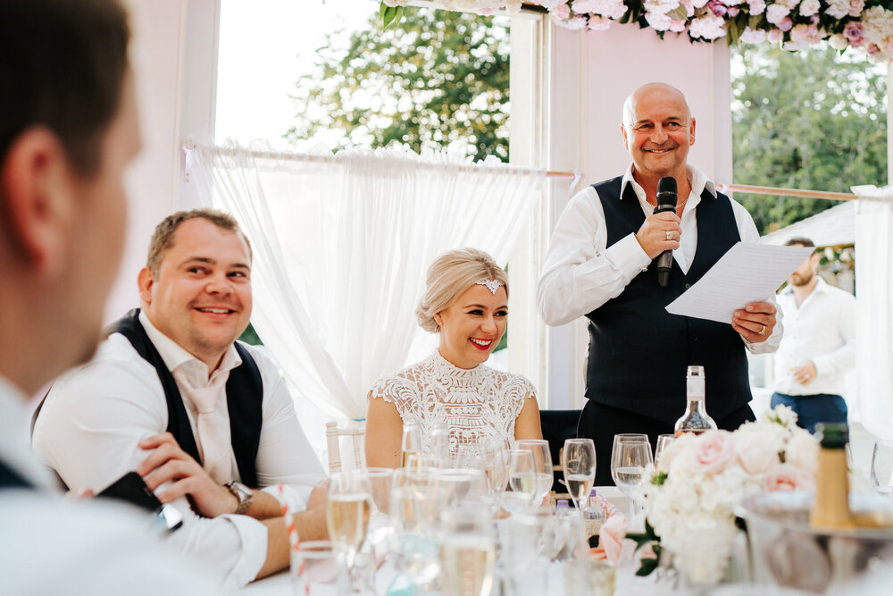 Father of the bride gives a speech as bride and guests react with smiles and laughter 