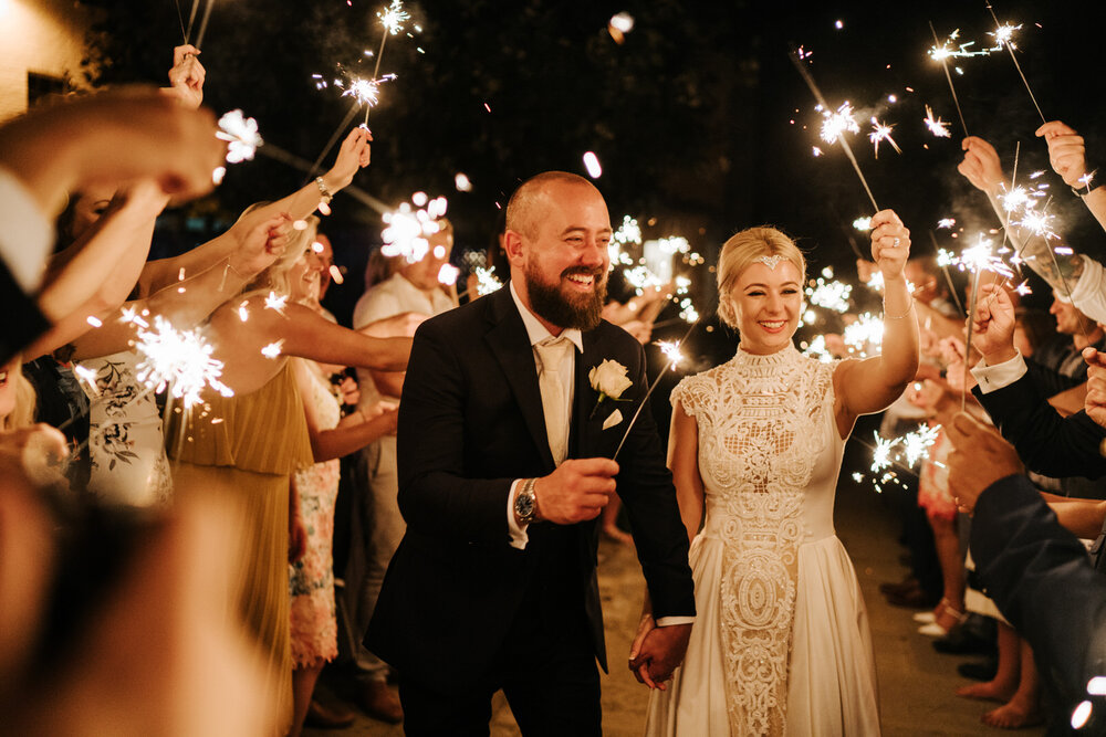 Guests form a tunnel of sparklers around the bride and groom as they hold sparklers and smile in excitement