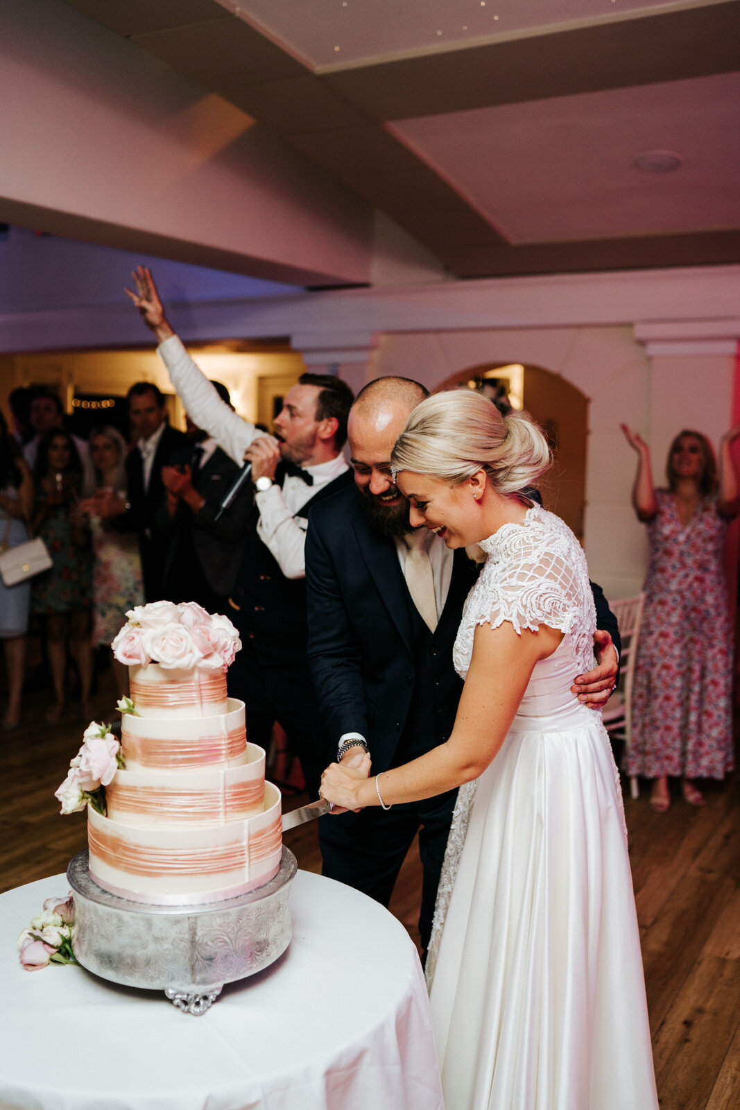 Bride and groom pose and cut the cake back inside at Pembroke Lodge as guests all around them cheer them on 