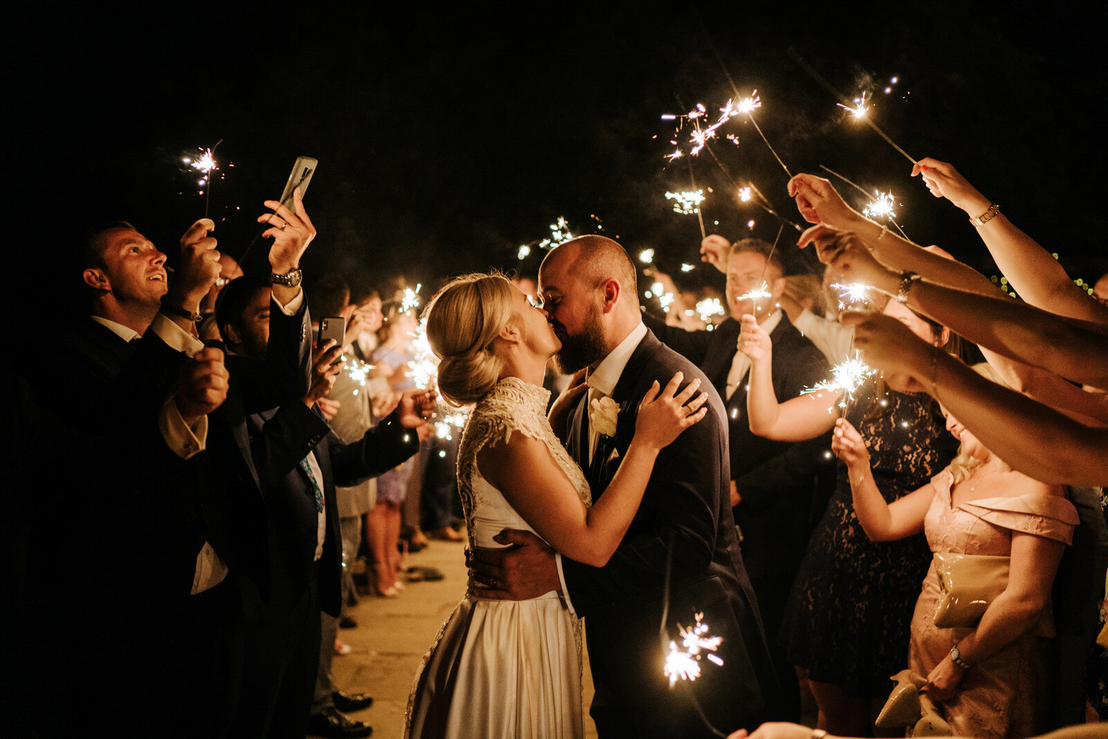 Guests form a tunnel of sparklers around the bride and groom as they hold sparklers and kiss in gorgeous framing outside at Pembroke Lodge during nighttime