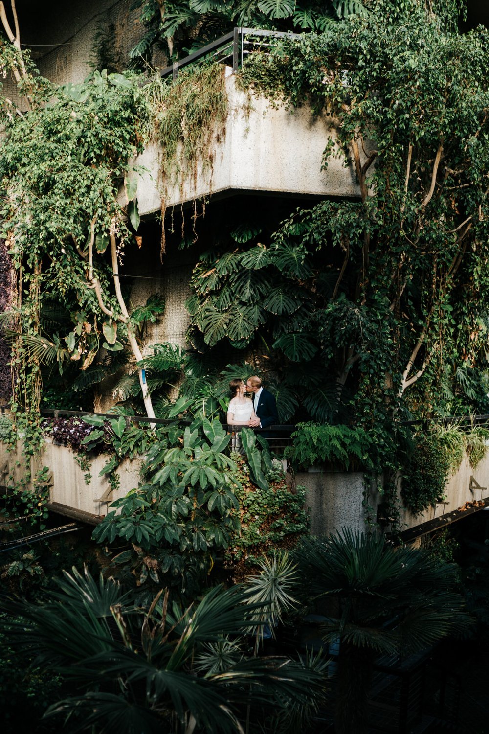 Bride and groom stand and kiss under beautiful green foliage at the Barbican Conservatory in London