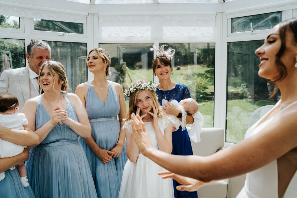 Bride reveals her dress to her bridesmaids and father while young flower girl looks at her in awe