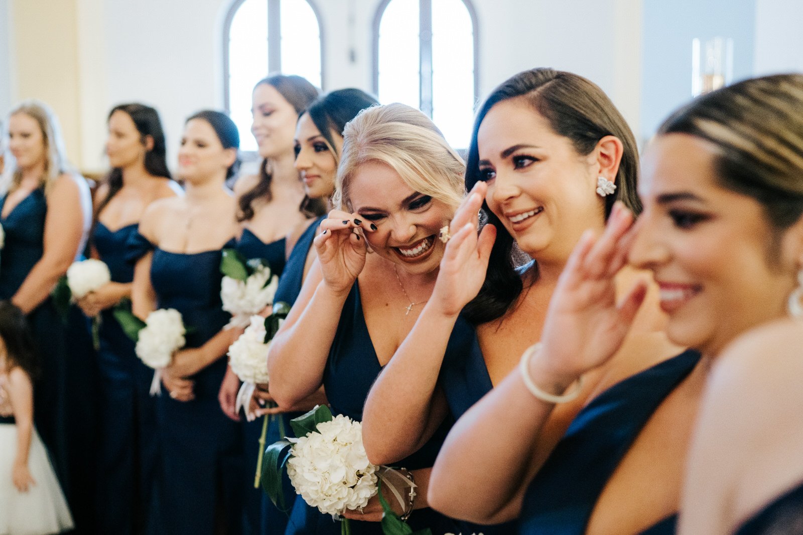 Eight bridesmaids wait at the top of the aisle of the church as bride walks down the aisle. Three of the bridesmaids wipe away tears of excitement