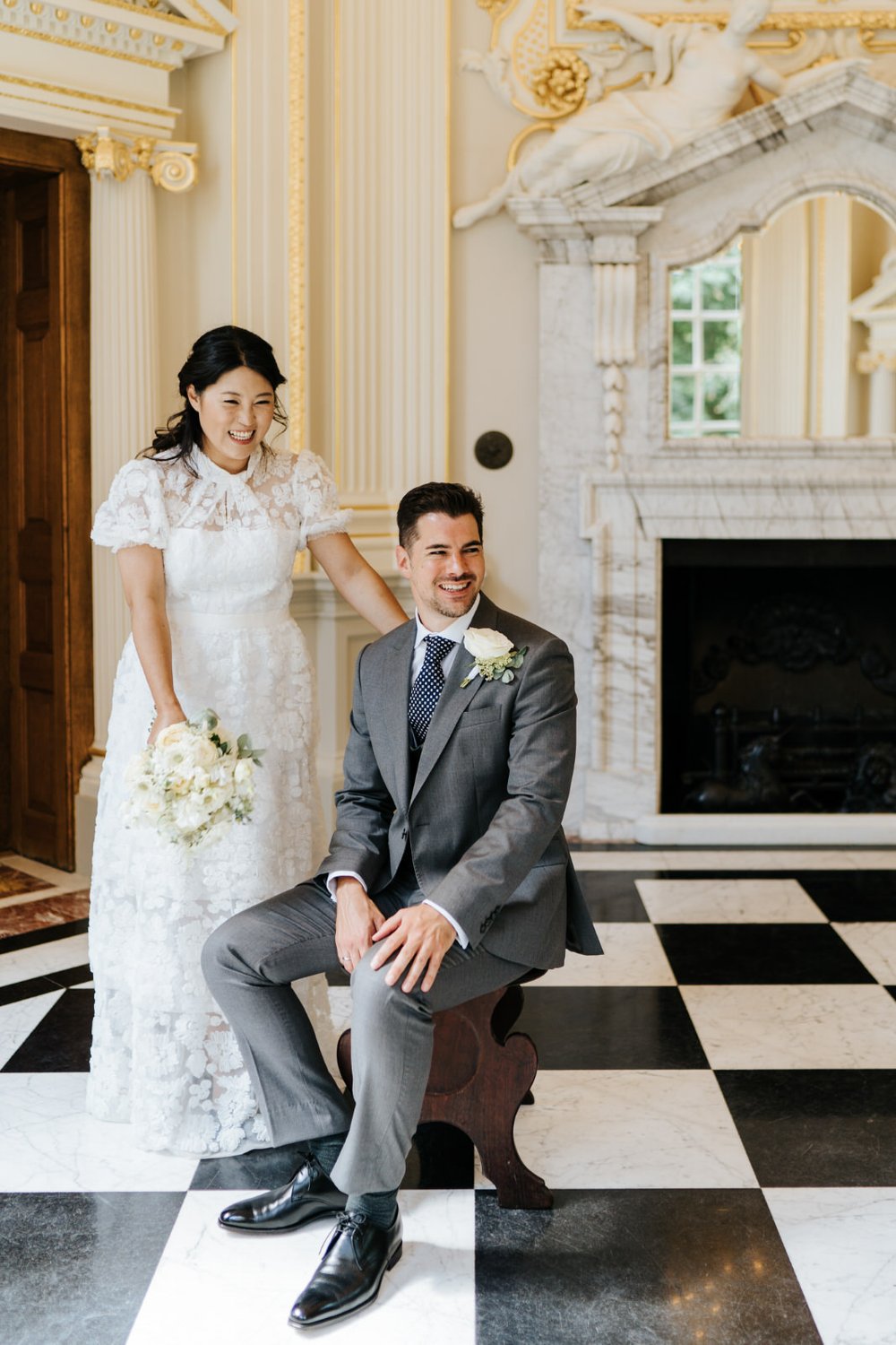 Groom sits on a chair as Korean bride stands next to him in wedding portrait at Orleans House Gallery
