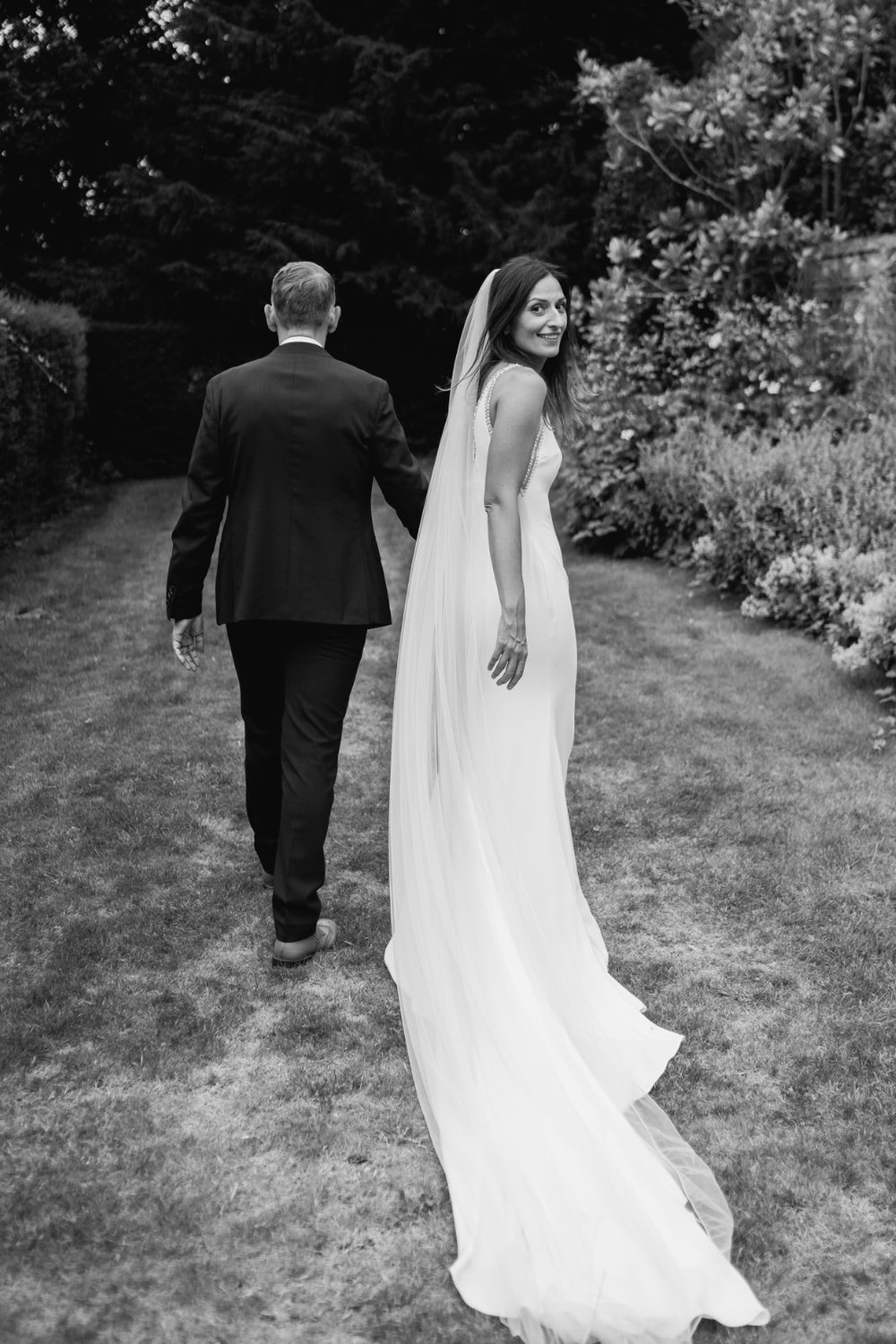 Bride and groom walk away from the camera as bride looks at camera over her shoulder with gorgeous long veiled dress at Foxhill Manor wedding