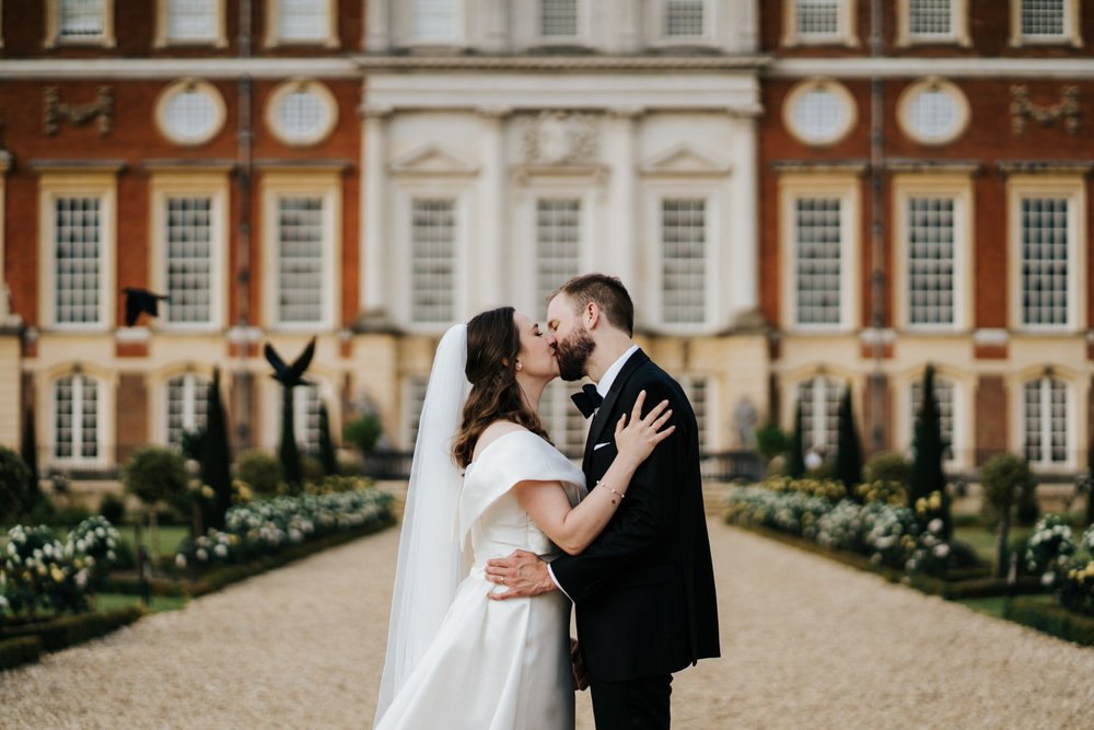Bride and groom pose in front of majestic Hampton Court Palace wedding backdrop as two birds fly on the left of the frame