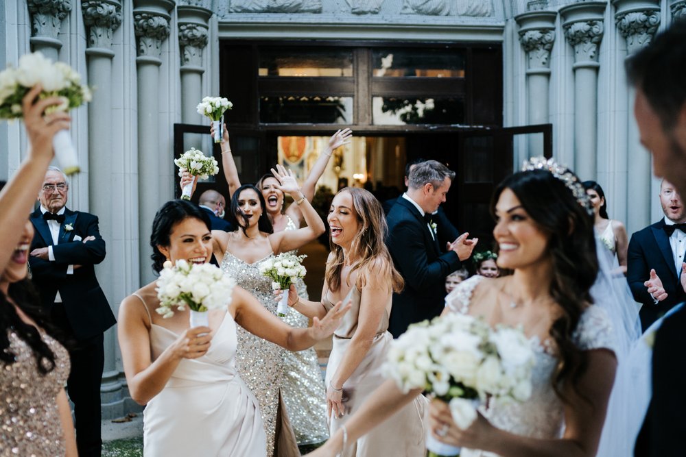 Bride, groom, bridesmaids and groomsmen jump and throw their hands into the air in unison cannot contain their excitement as they exit the church after wedding ceremony