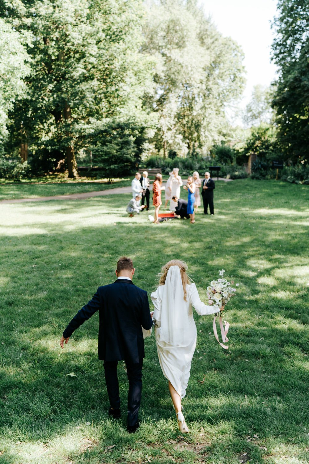Bride and groom, hand in hand, walk down a slope to join their wedding guests on the lawn of Orleans House Gallery 