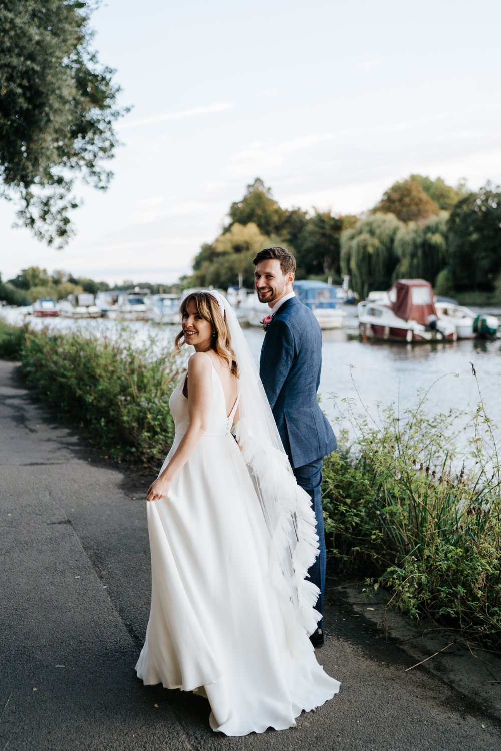 Bride and groom walk away from camera and look towards it over their shoulder during photo session on Richmond Riverside