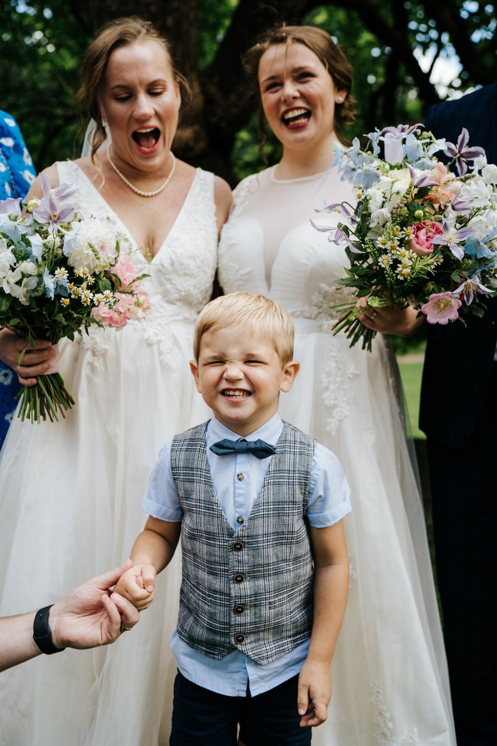 Young boy smiles aggressively as both brides stand behind him during family portraits