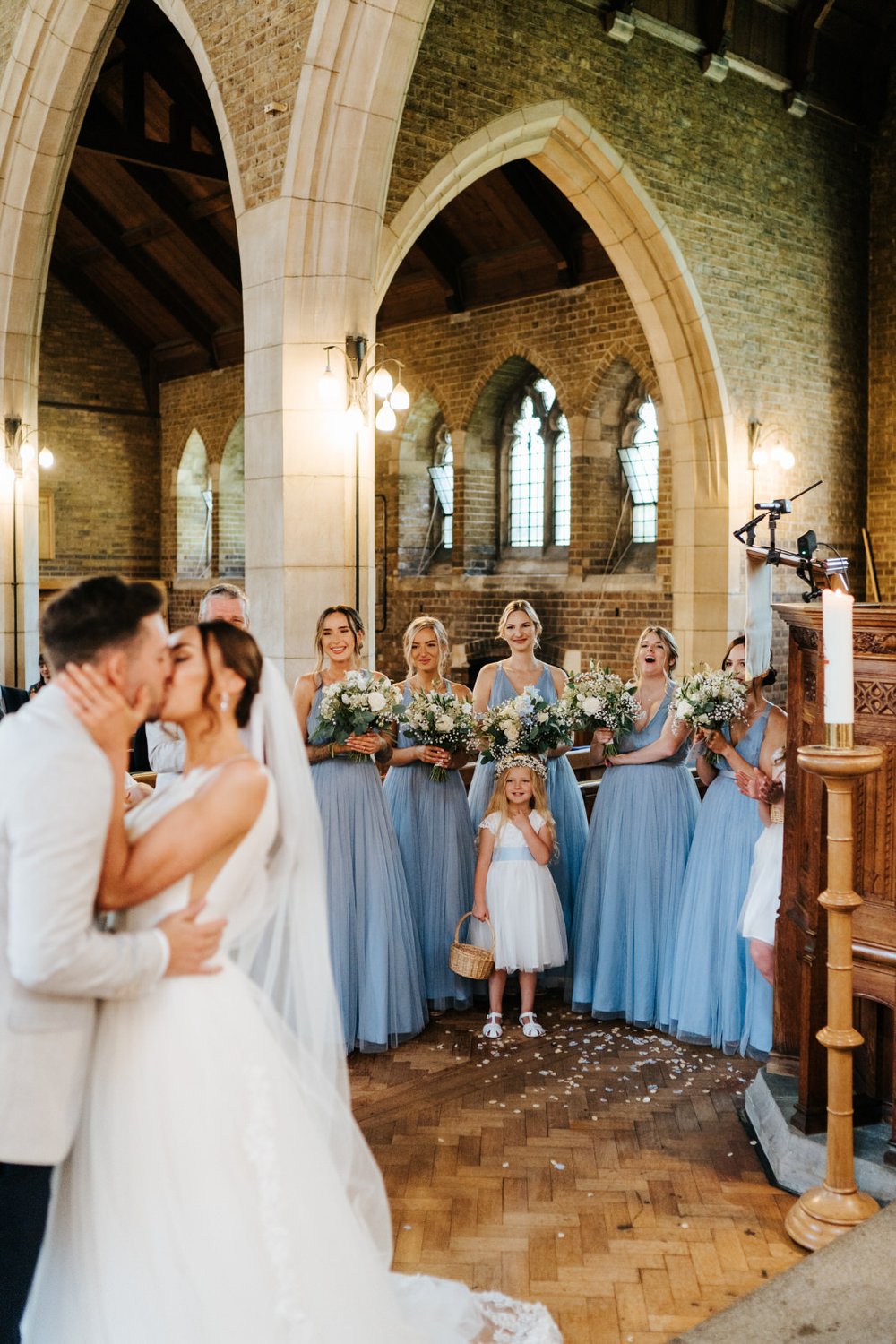 Photograph taken from the altar of the bride and groom, out of focus, sharing their first kiss while bridesmaids smile in the background