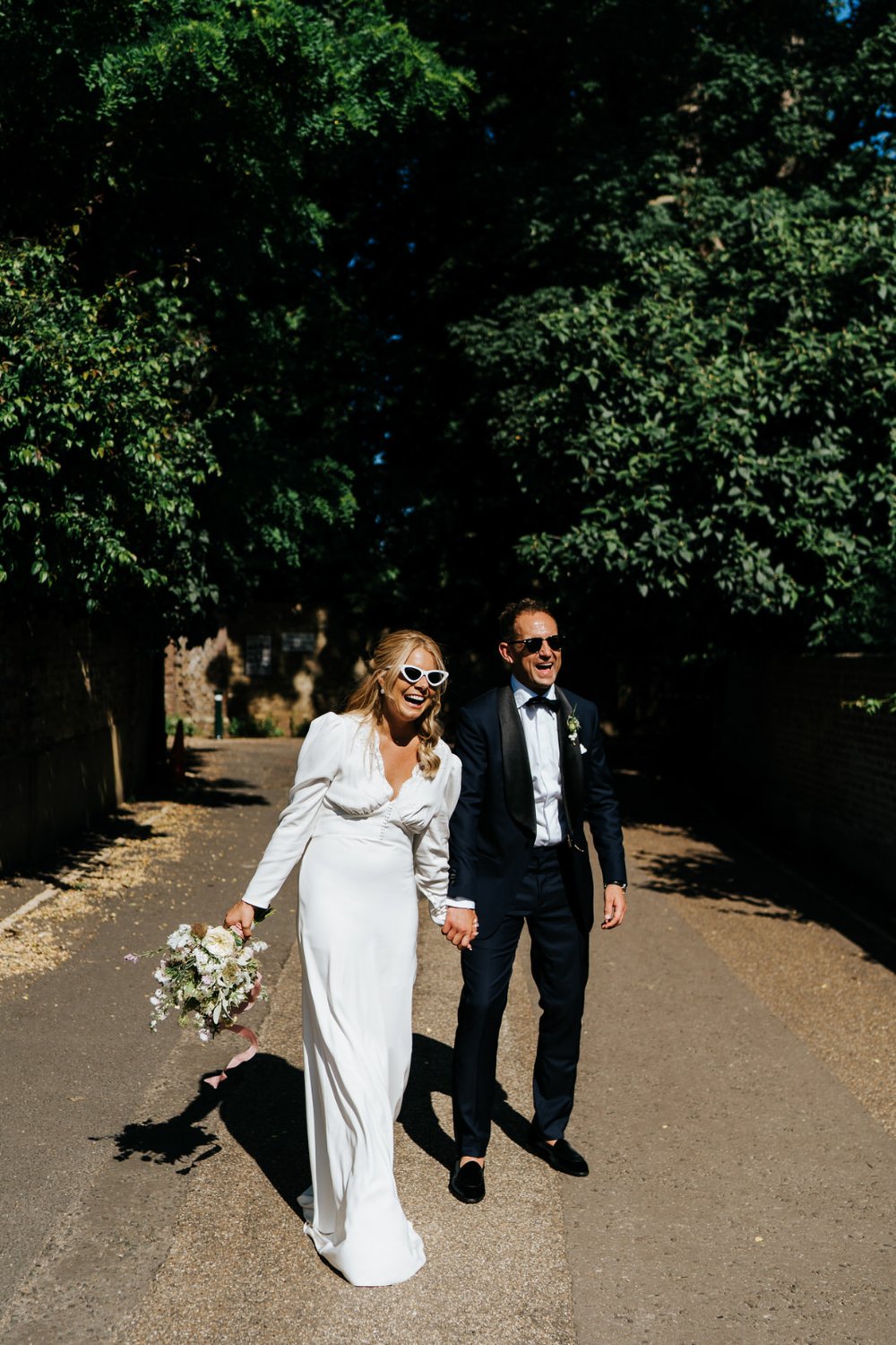Bride and groom, basking in the sun, smile as they look dapper in wedding outfit and sunglasses