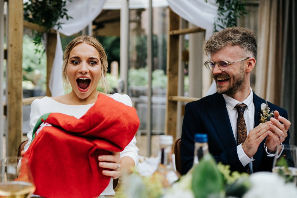 Bride, sitting next to groom, receives unexpected gift during wedding speeches at Hampton Manor in Arden wedding and reacts with utter disbelief